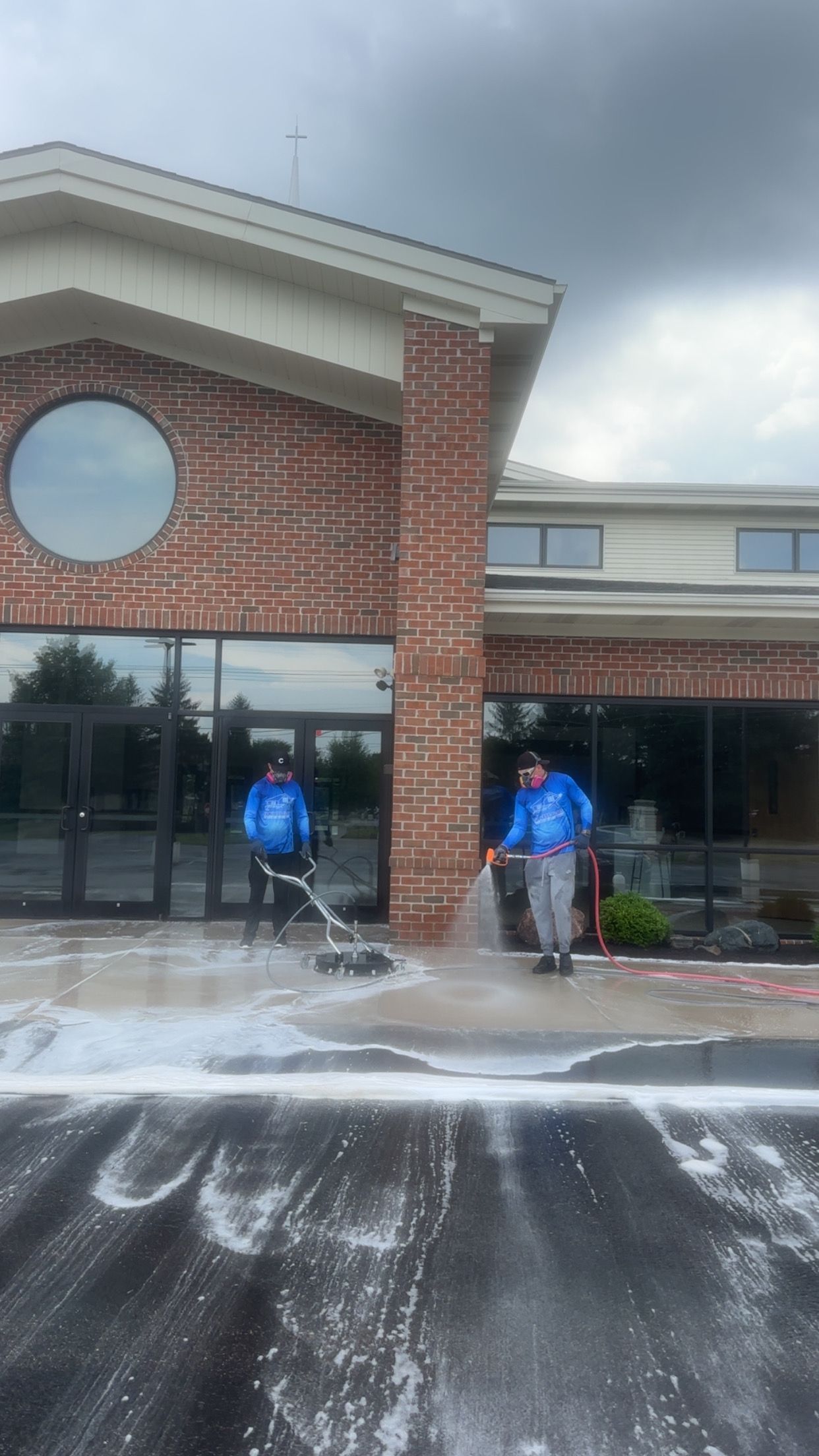 Two people in blue shirts standing by a brick building with water spraying across the driveway