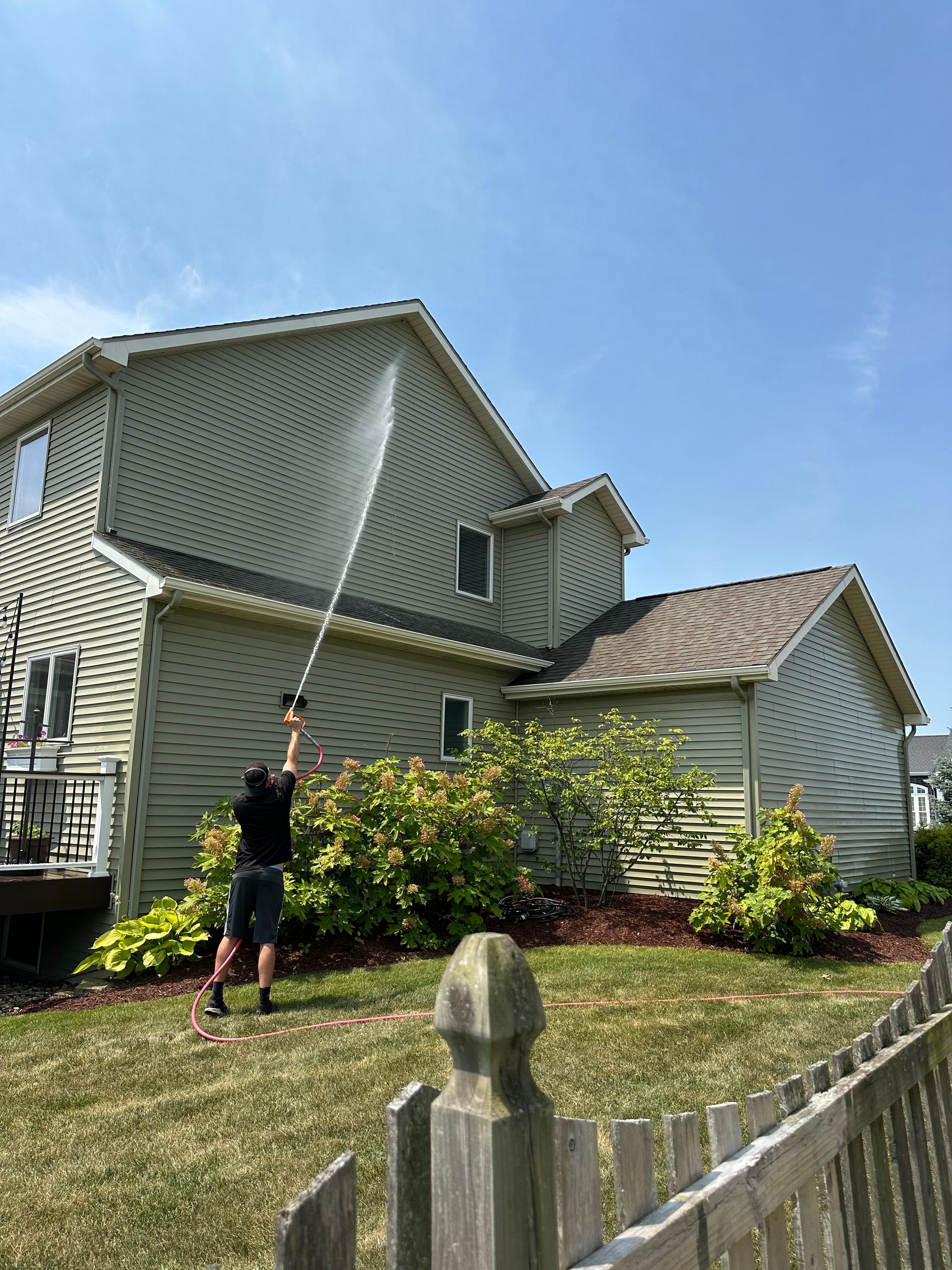 Person spraying water onto a house with a hose in a sunny yard