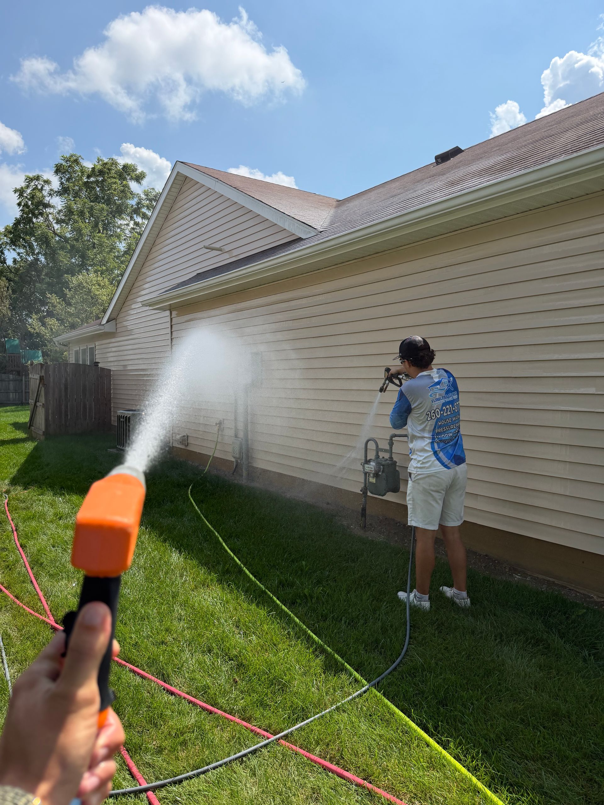 Person sprays a house wall with a garden hose nozzle in a sunny backyard.
