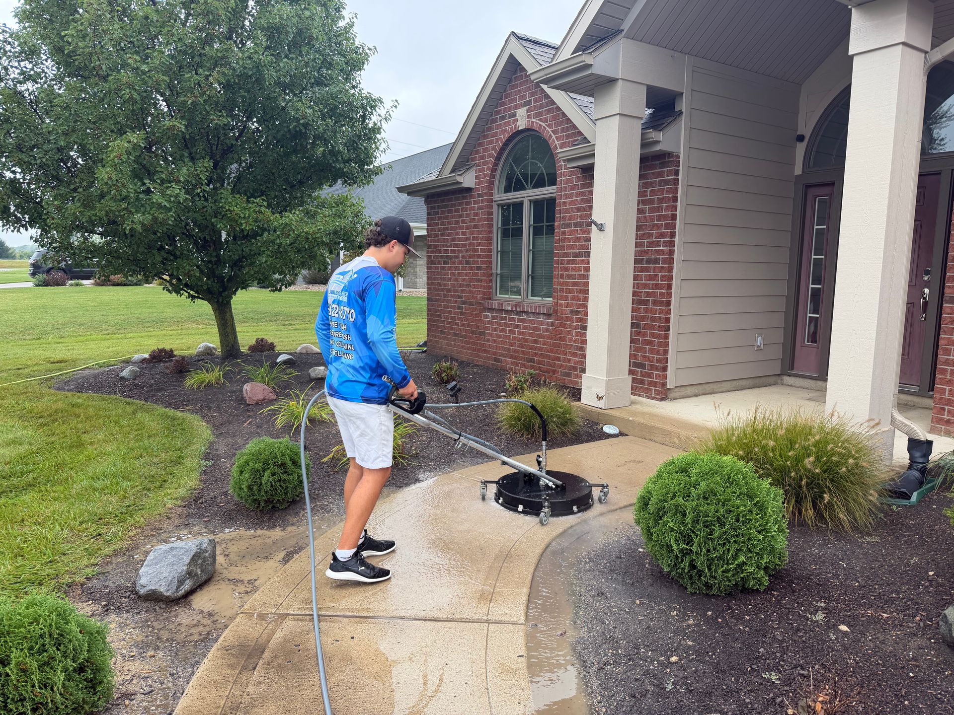 Person watering small shrubs along a sidewalk beside a brick house