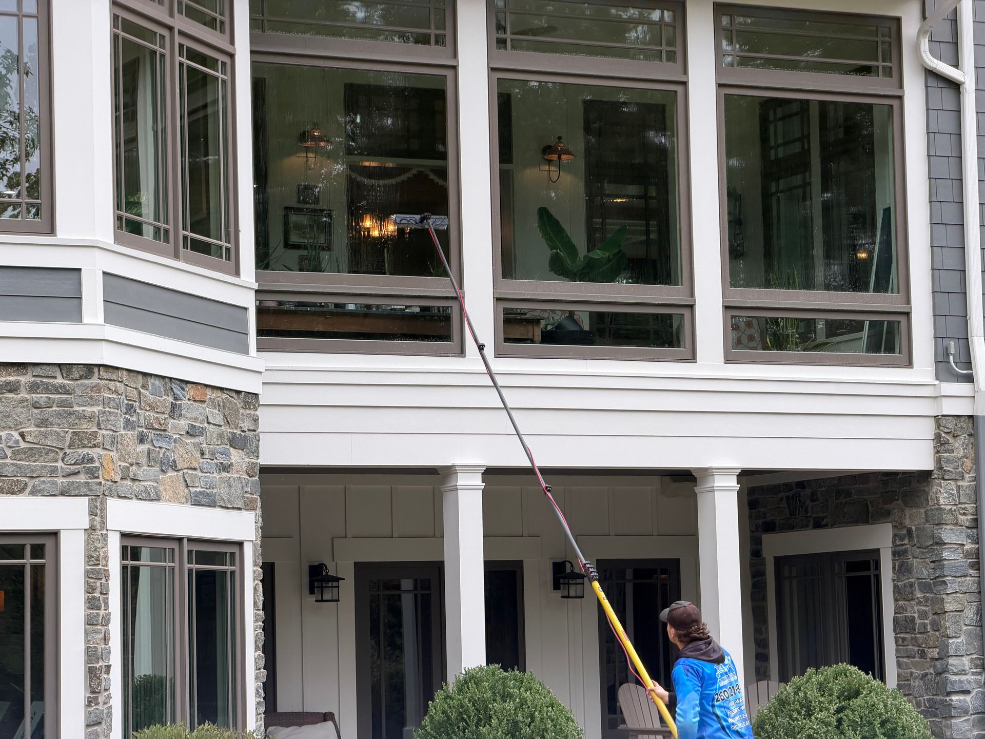 Person on a ladder uses a long brush to clean upper windows of a house.