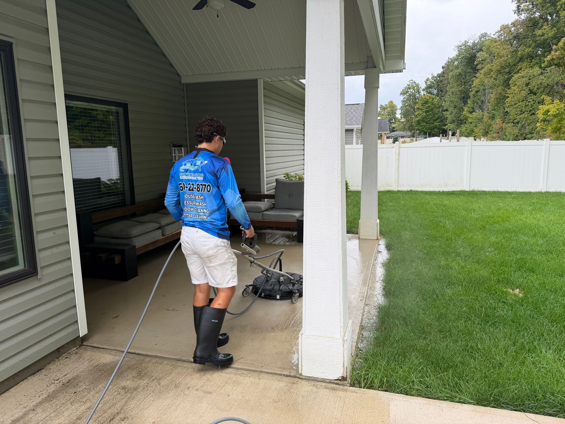 Person power washing a covered patio beside a green lawn and white fence.