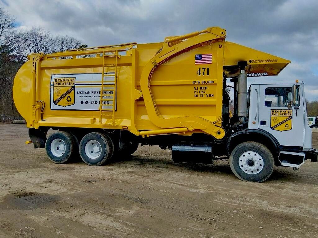 A yellow sanitation truck parked on a gravel lot under a clear blue sky, with trees in the background.