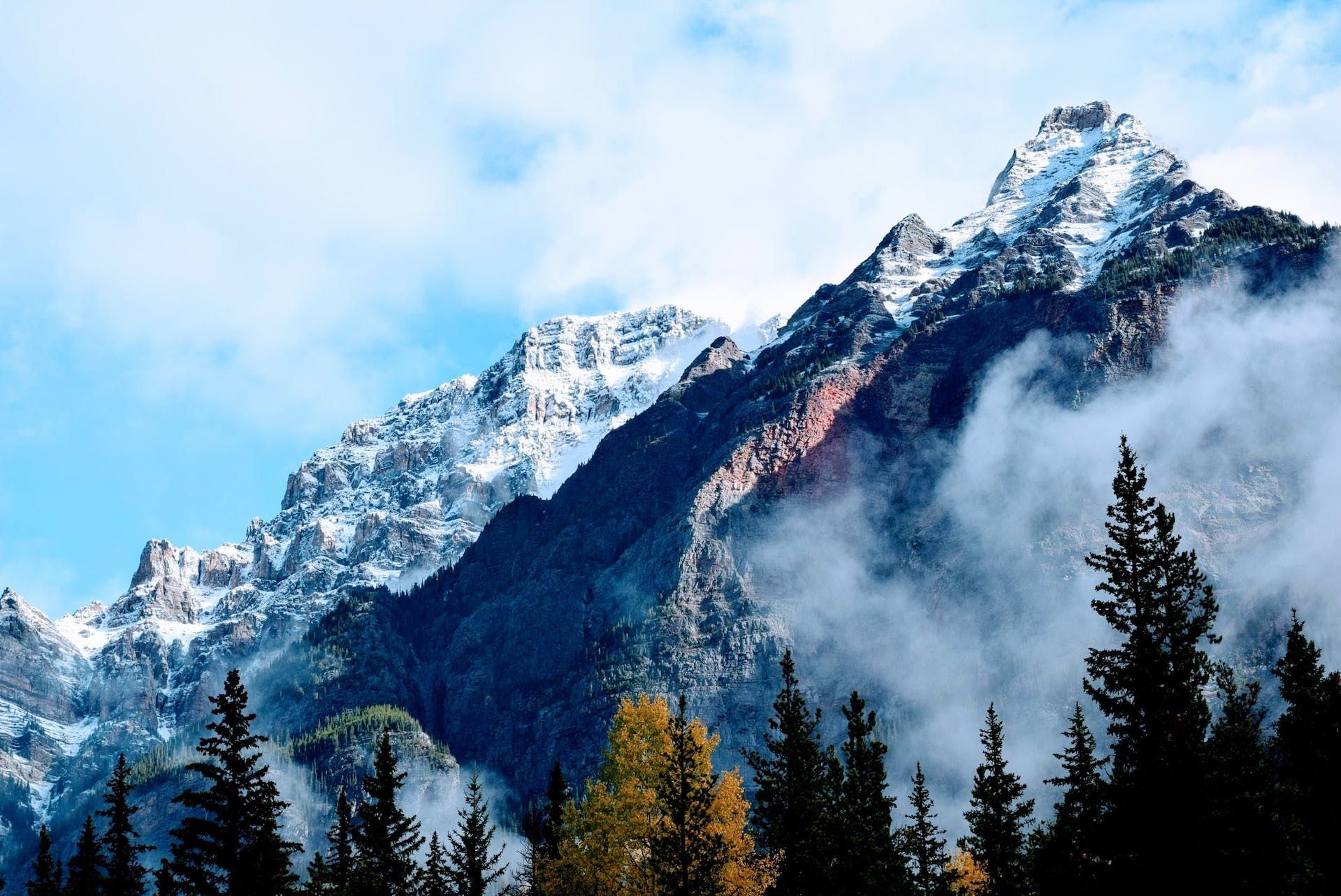 A mountain covered in snow and clouds with trees in the foreground
