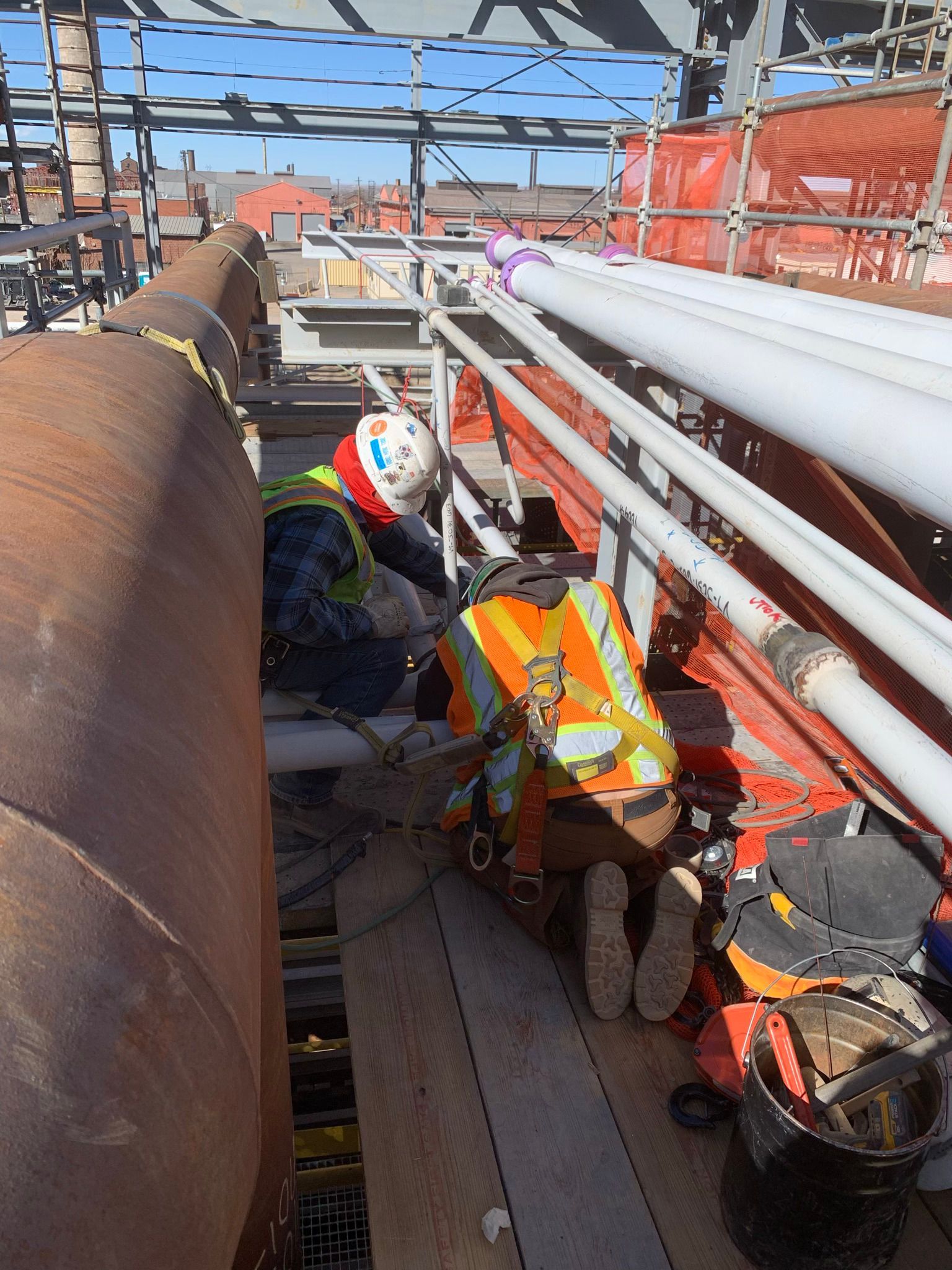 Two construction workers are working on a pipe on a scaffolding.