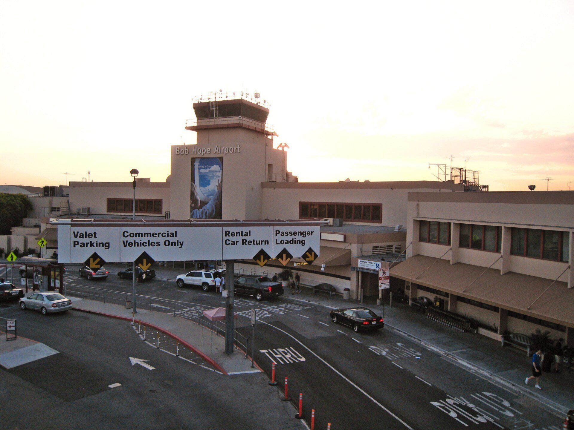 Exterior view of the terminal at Hollywood Burbank Airport, with cars entering and exiting the passenger loading area. The sky is a blend of orange and pink.