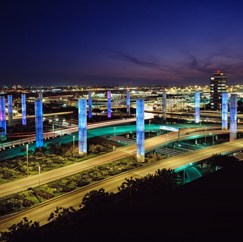 Night view of Los Angeles with illuminated blue and purple pylons over a freeway. Tall buildings and a dark sky are visible in the background.