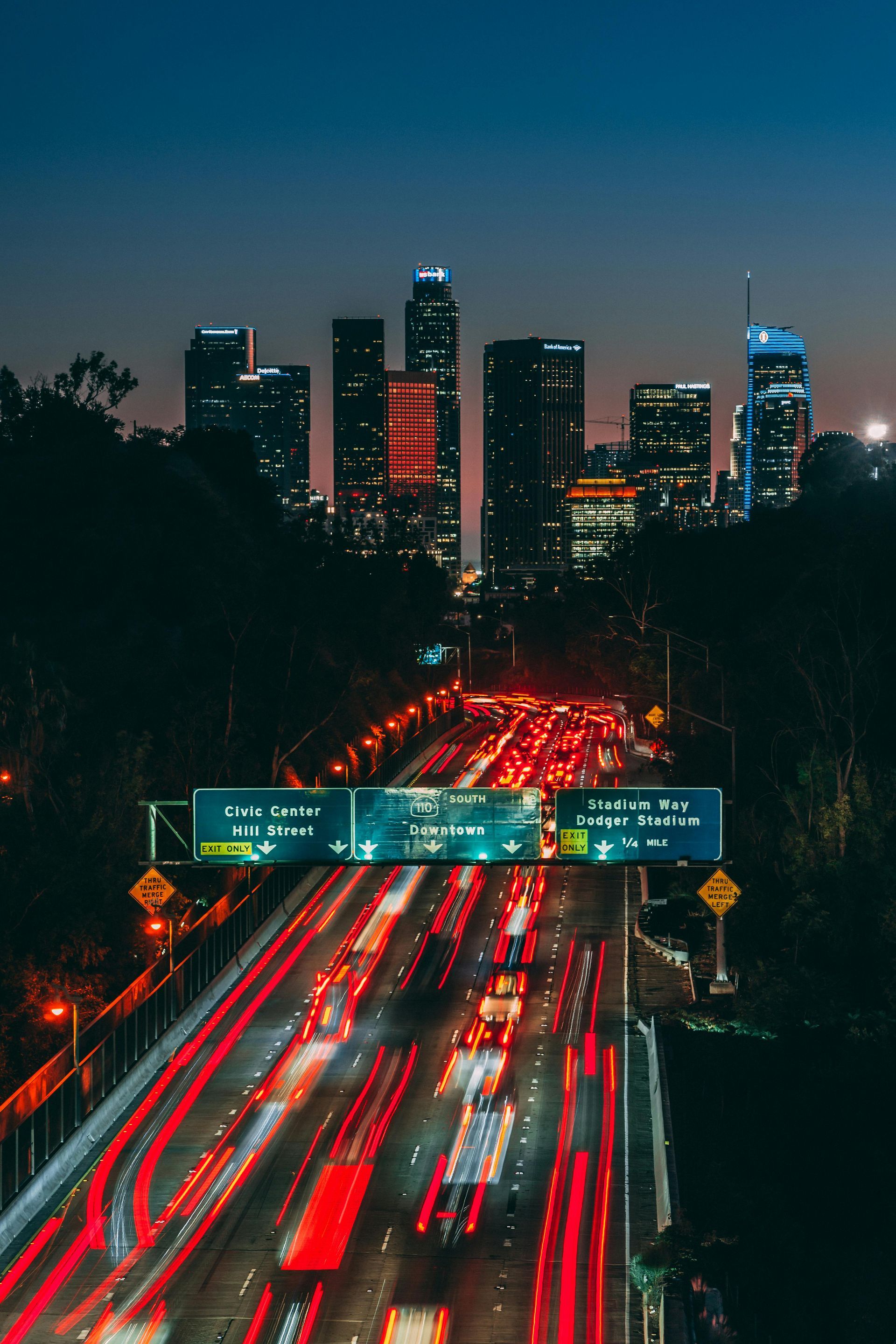 Nighttime view of a highway filled with streaks of red car lights, leading to a distant city skyline with illuminated buildings.