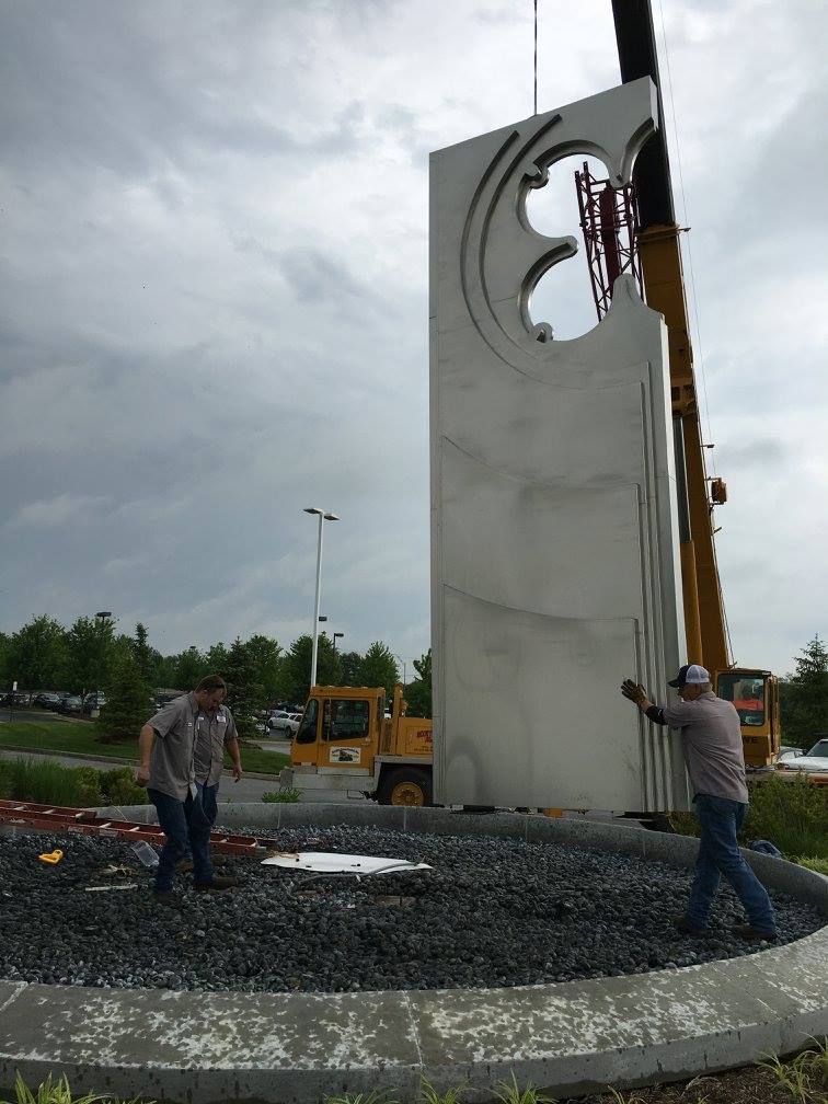 Workers inspect a tall white sculpture on a gravel roundabout under a cloudy sky
