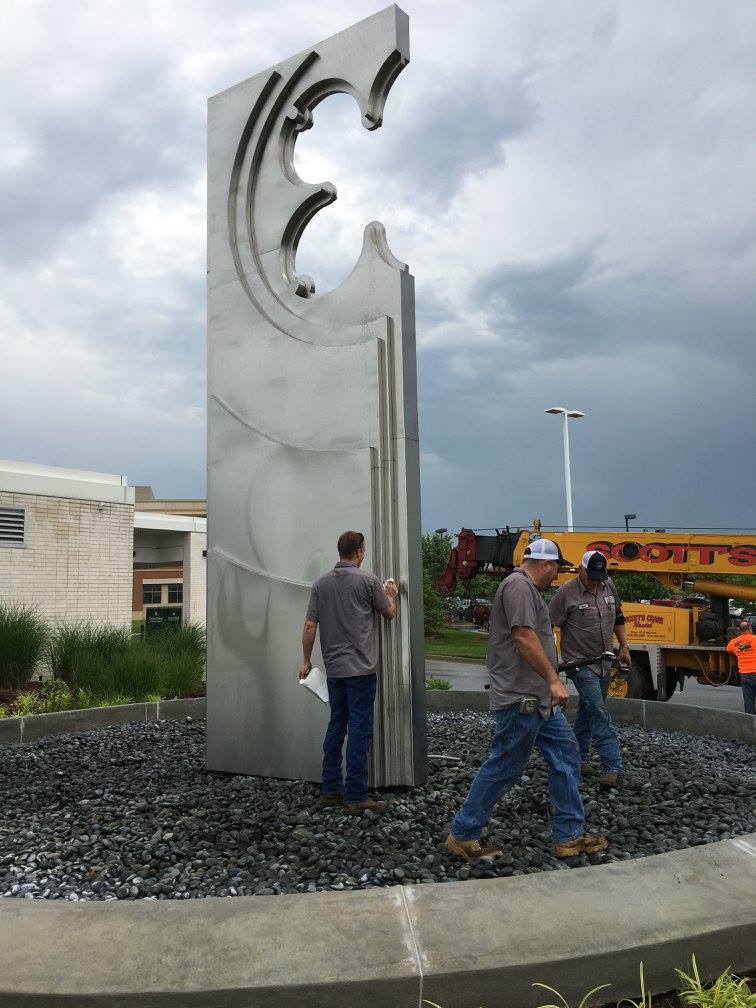 People viewing a tall silver abstract sculpture on a gravel base outdoors under cloudy skies