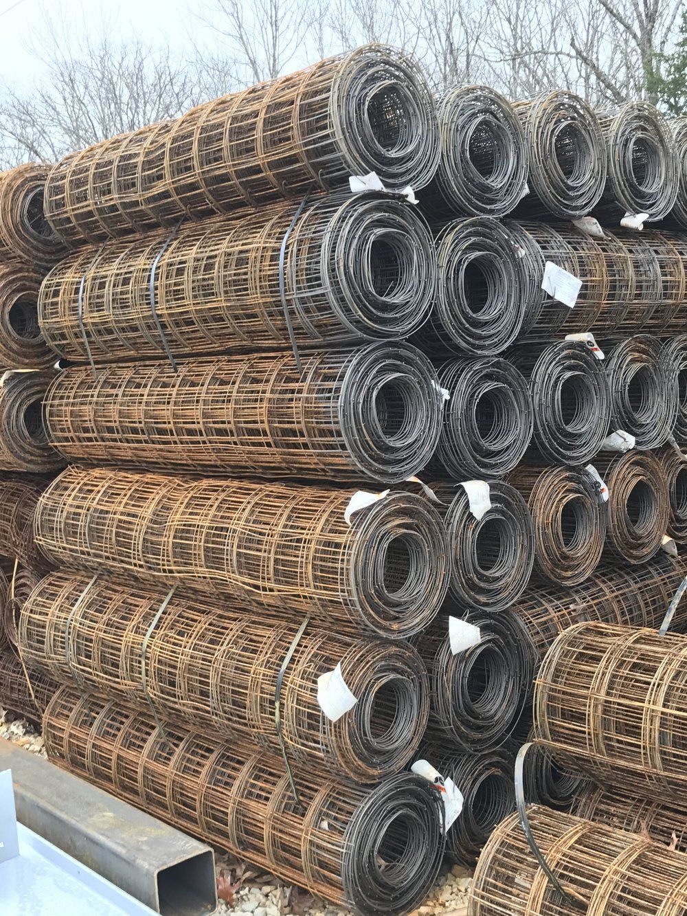 Stacks of rolled wire mesh sheets outdoors against a bare tree backdrop.