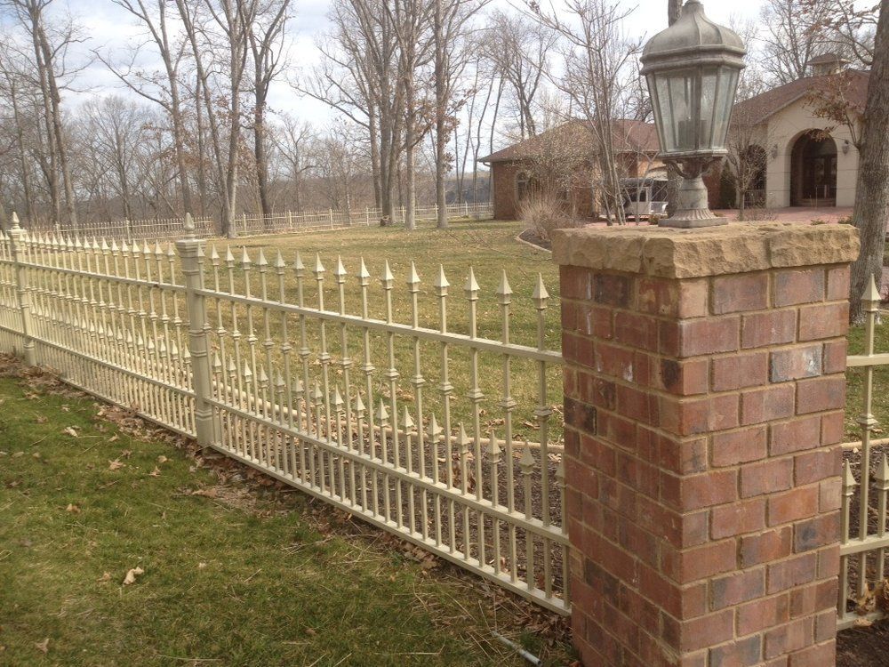 Brick pillar and cream metal fence beside a lawn, with a house and bare trees in the background