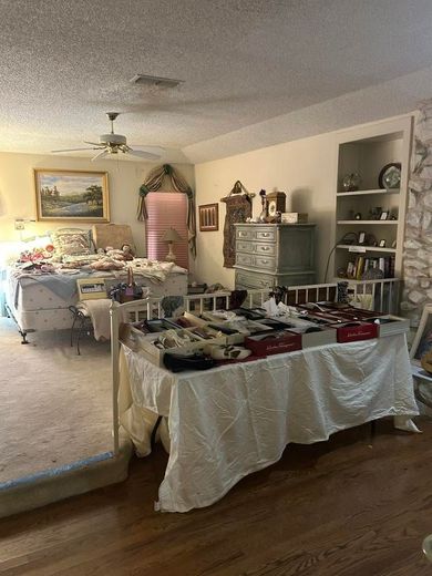 Cluttered bedroom with two beds, dresser, mirror, and laundry on a table in warm indoor light