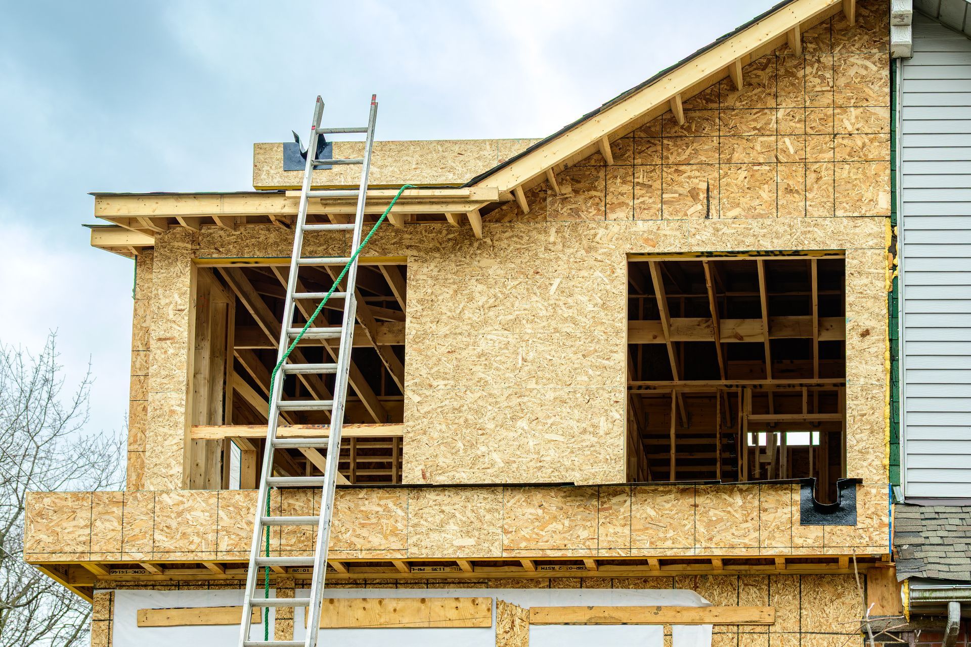 Maison en construction avec charpente en bois apparente, échelle et ciel nuageux.