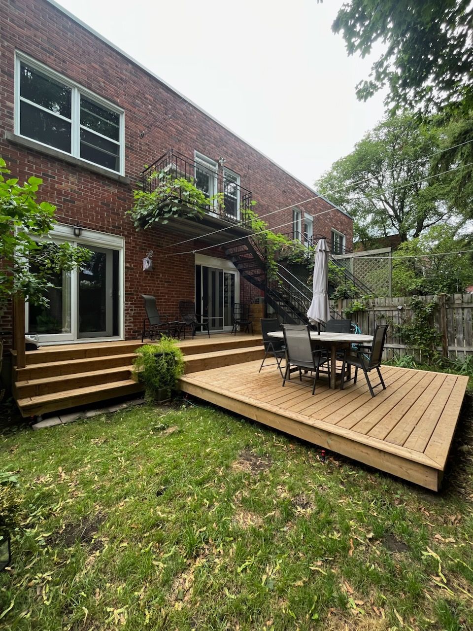 Une terrasse en bois prolonge un bâtiment en briques doté d'un escalier de secours en fer forgé. Une table, des chaises et un parasol sont installés sur la terrasse.
