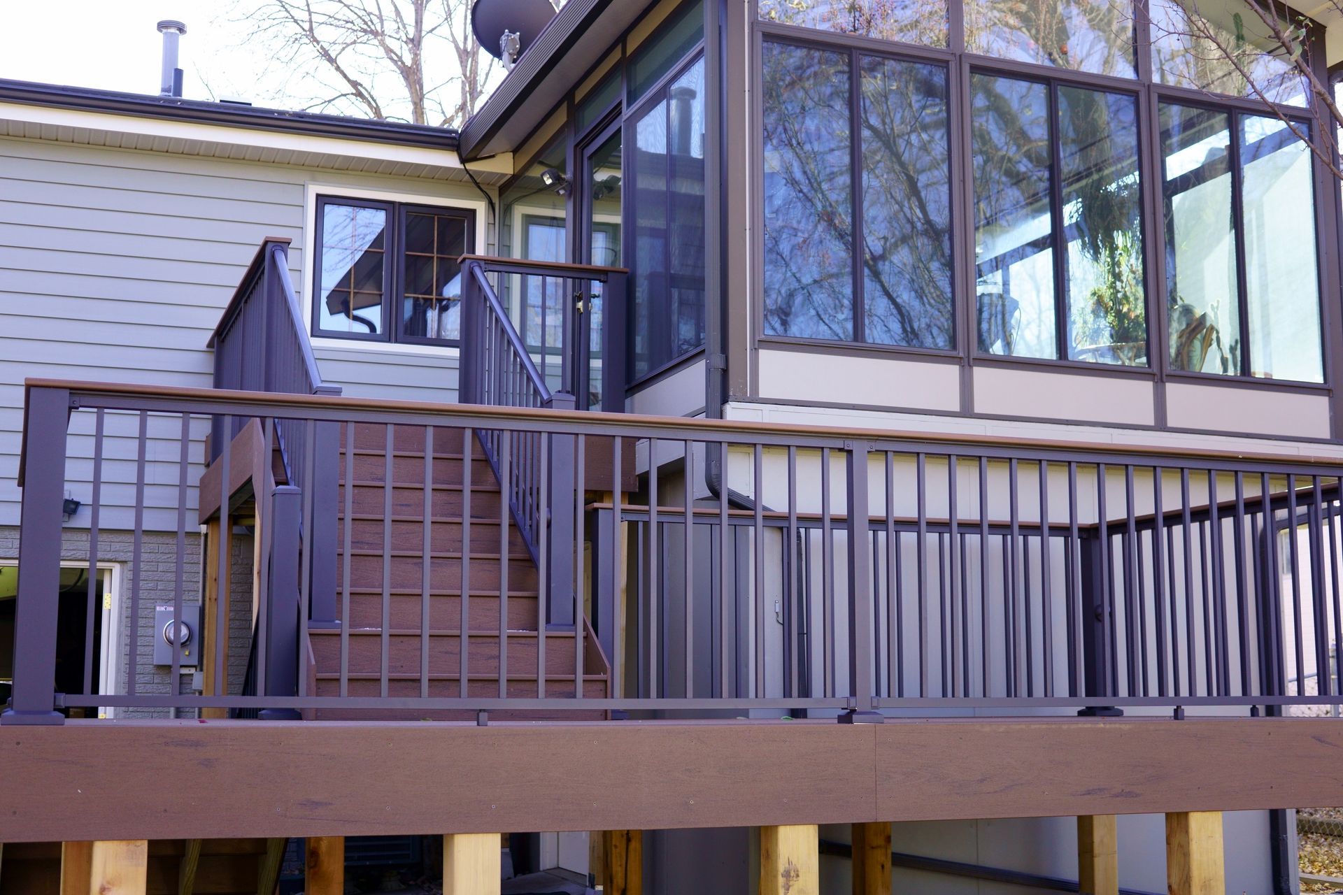 Terrasse avec rambarde en métal, escalier et véranda attenante avec de grandes fenêtres reflétant les arbres.