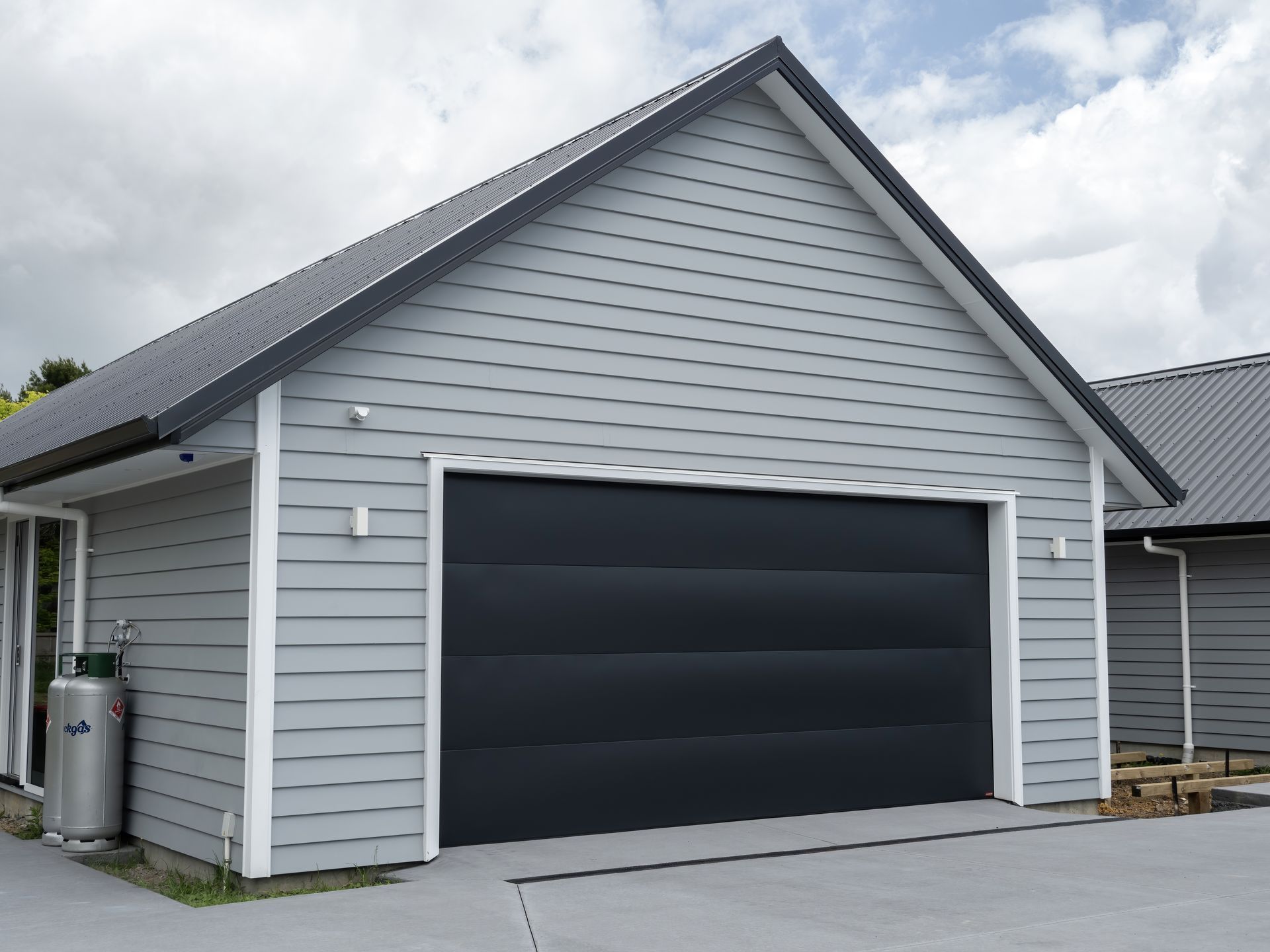 Garage gris avec porte, bardage et toit noirs sous un ciel nuageux.