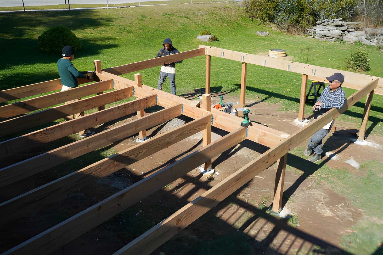 People assembling a wooden deck frame outdoors with tools and beams visible