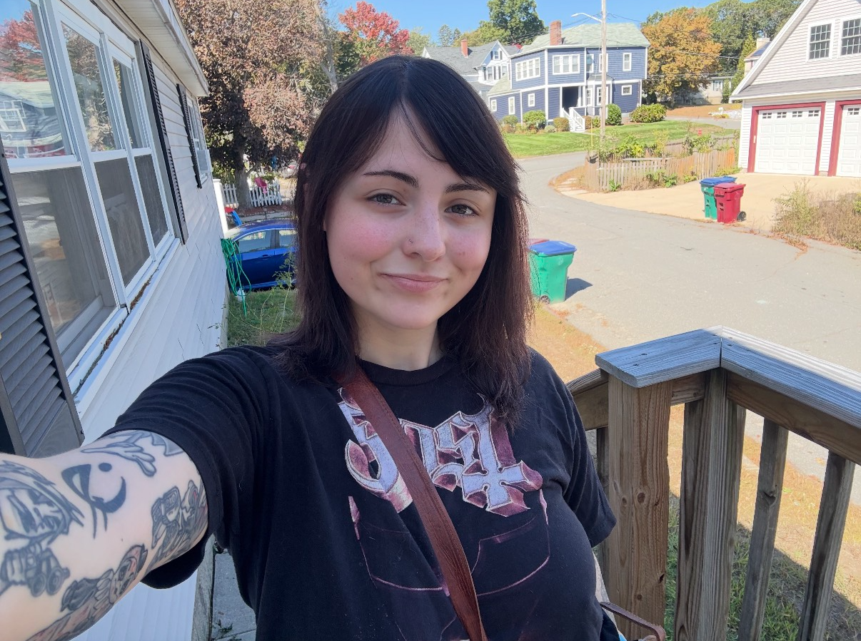Person with arm tattoos smiles outdoors, taking a selfie. Houses and a tree-lined street in the background.