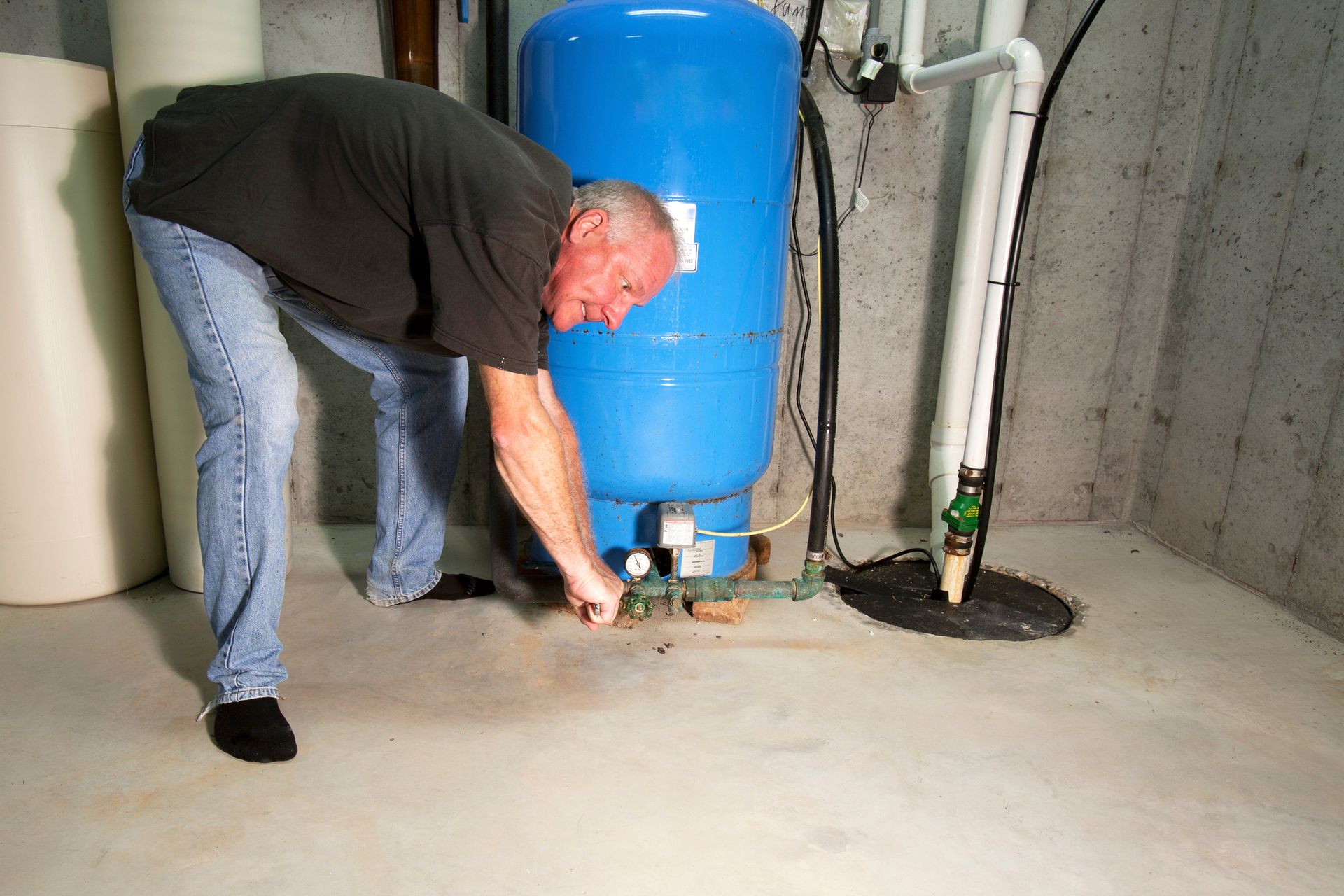 Man working on plumbing near a blue water tank in a basement, concrete floor.