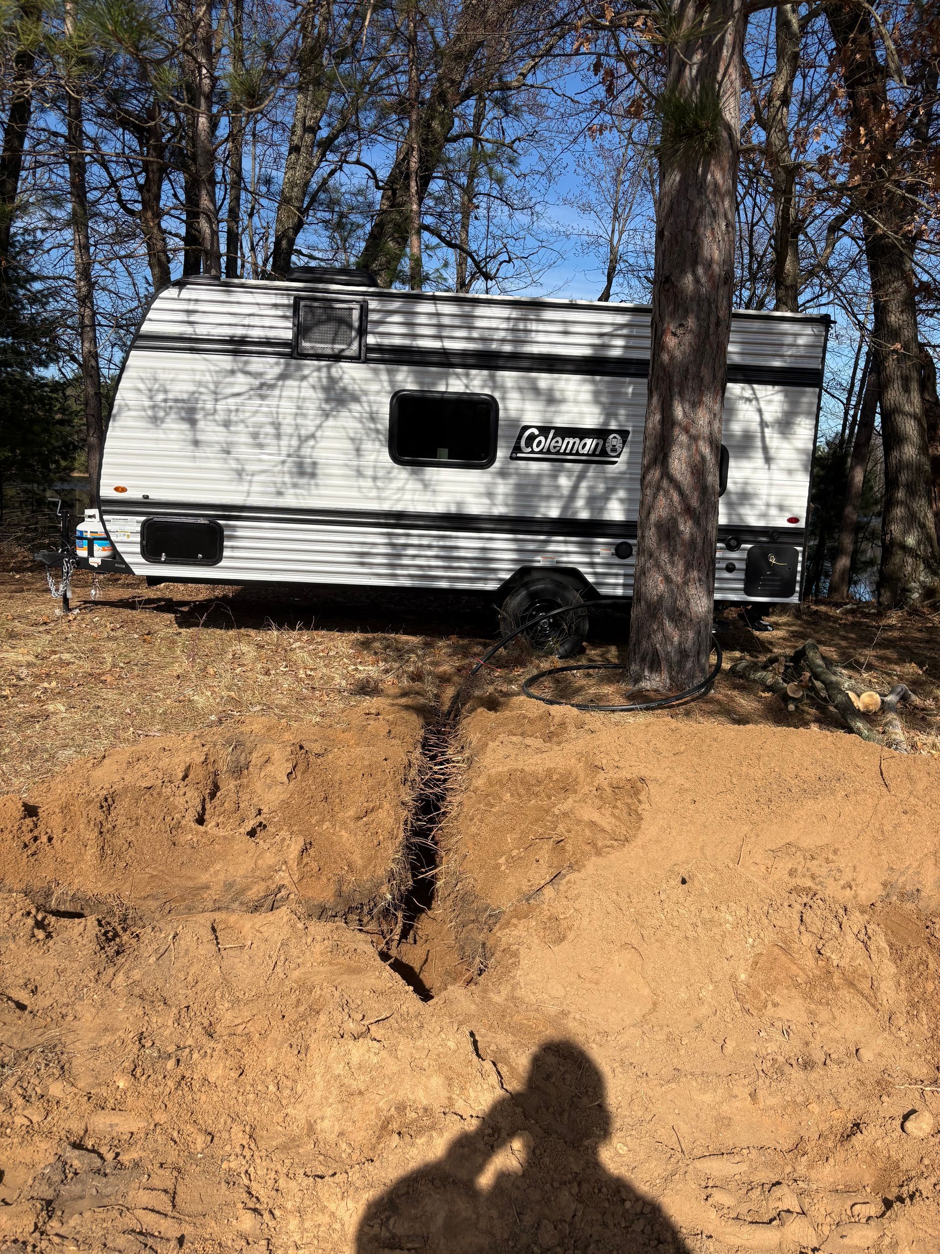 White camper trailer parked in woods beside a dirt embankment, with tree shadows and a blue sky.