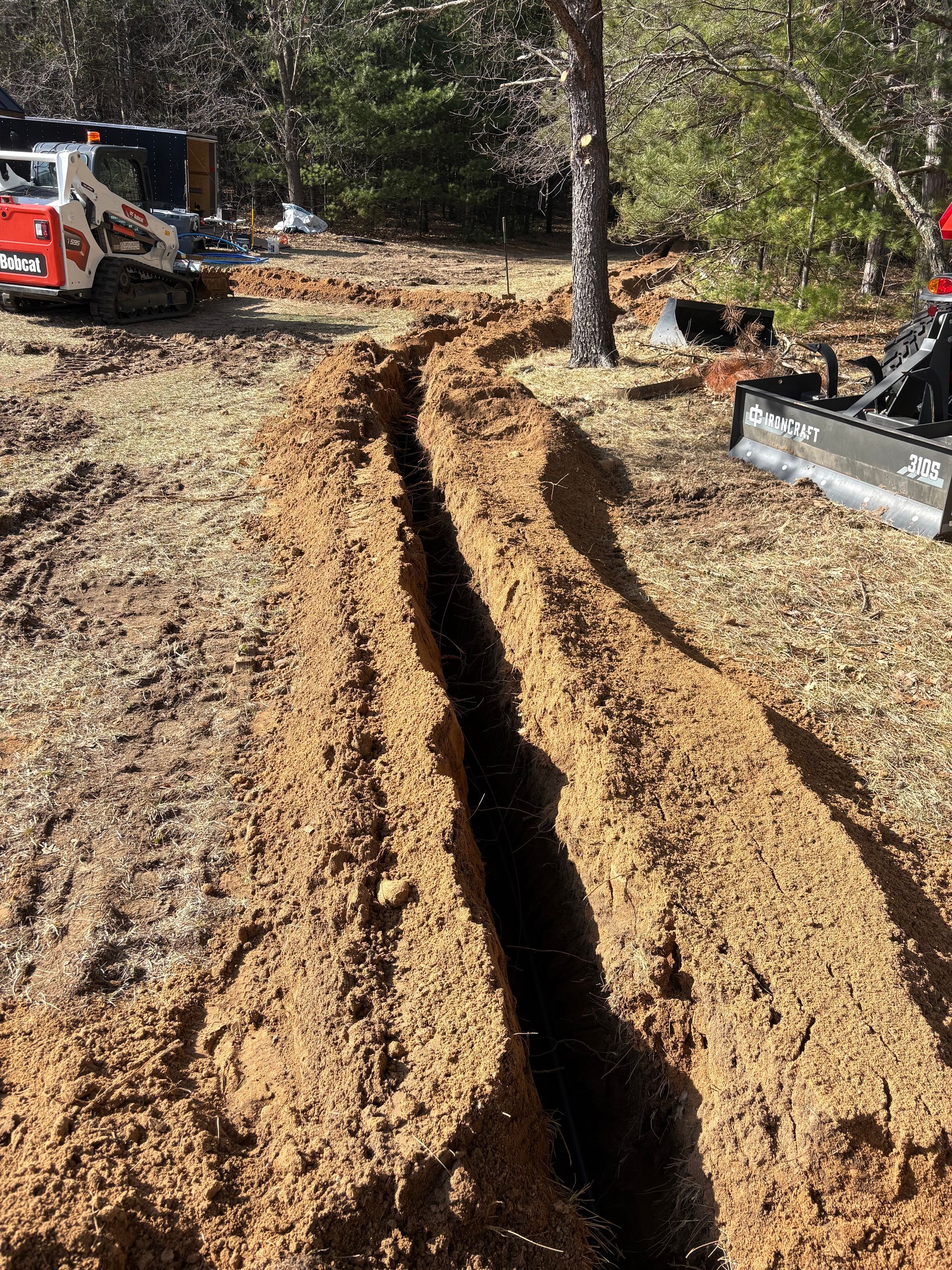 Deep trench cut through muddy ground at a construction site with an excavator nearby.