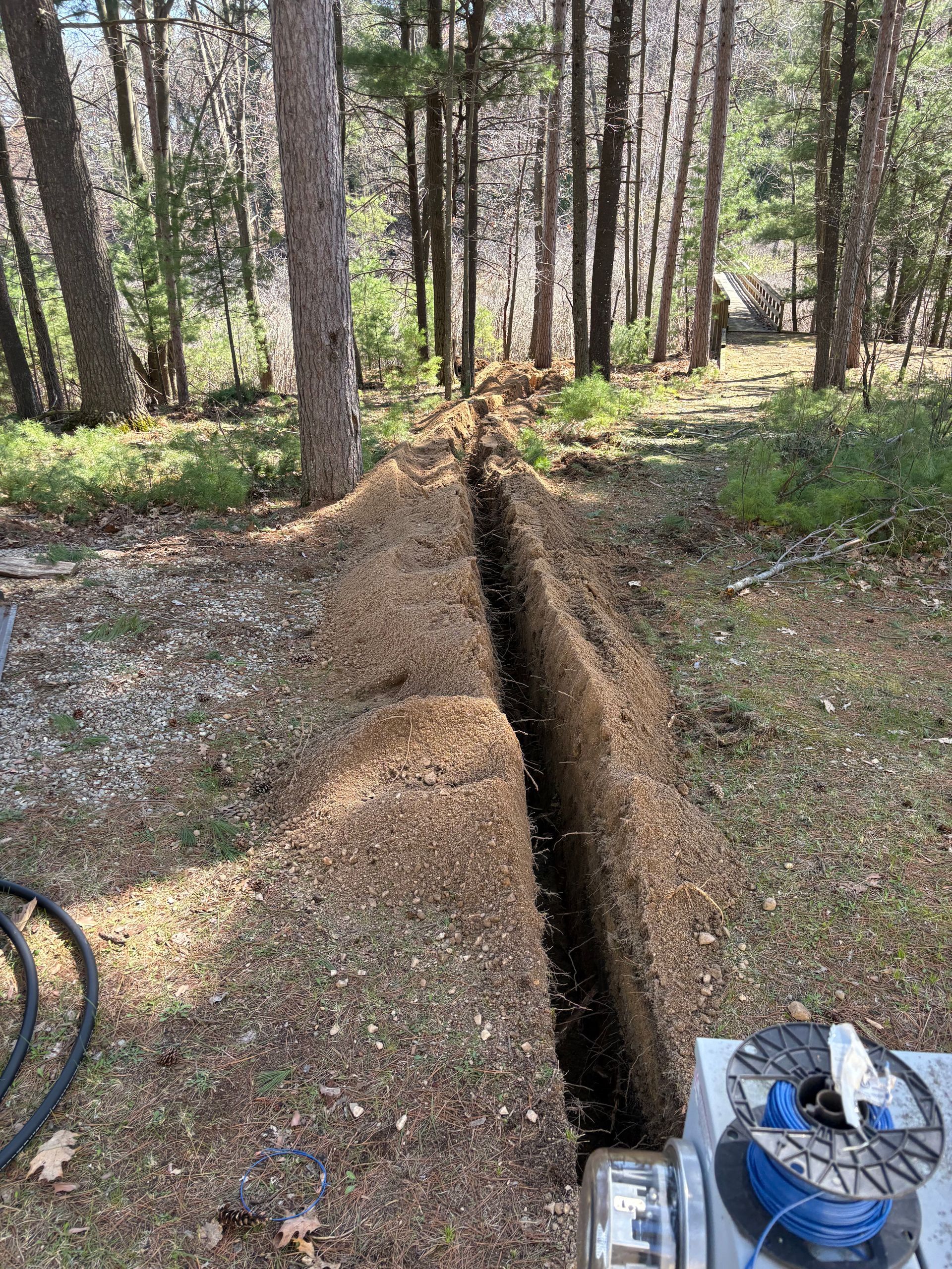 Forest clearing with a narrow trench cut through gravel and soil, trees in the background, and equipment at the edge