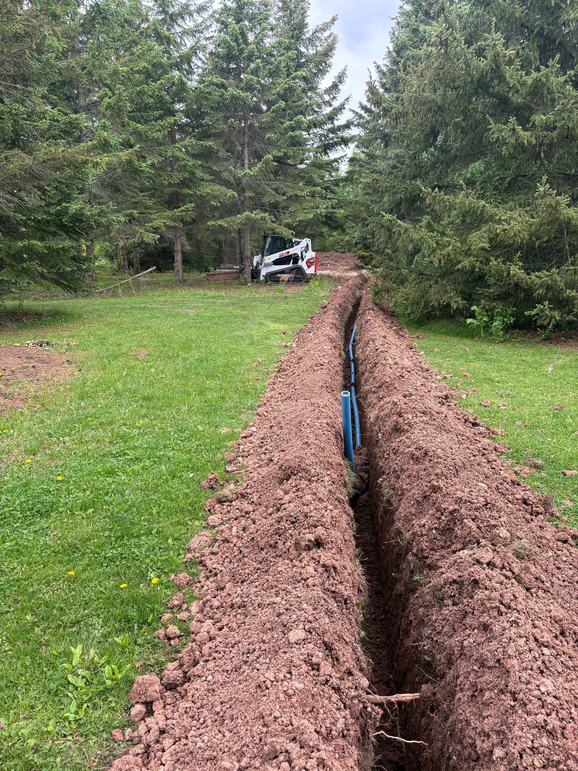 A long trench cut through a grassy yard, with a blue pipe visible and a small excavator in the distance.