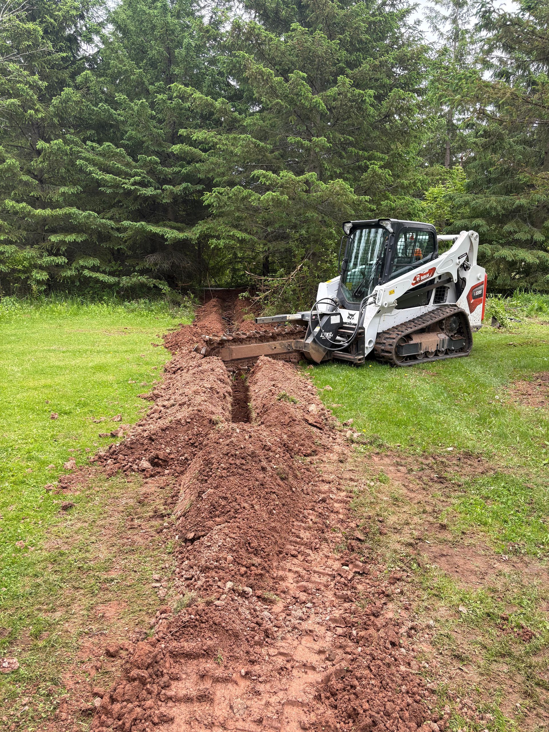 White compact excavator digging a muddy trench through grassy ground beside trees