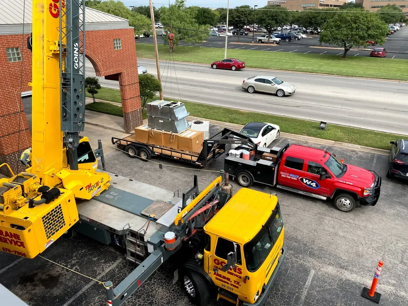 Crane and trucks moving equipment beside a brick building on a city street