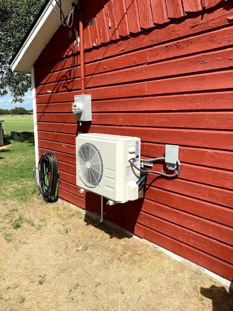 Outdoor HVAC units mounted on a red barn wall beside a coiled hose in a grassy yard