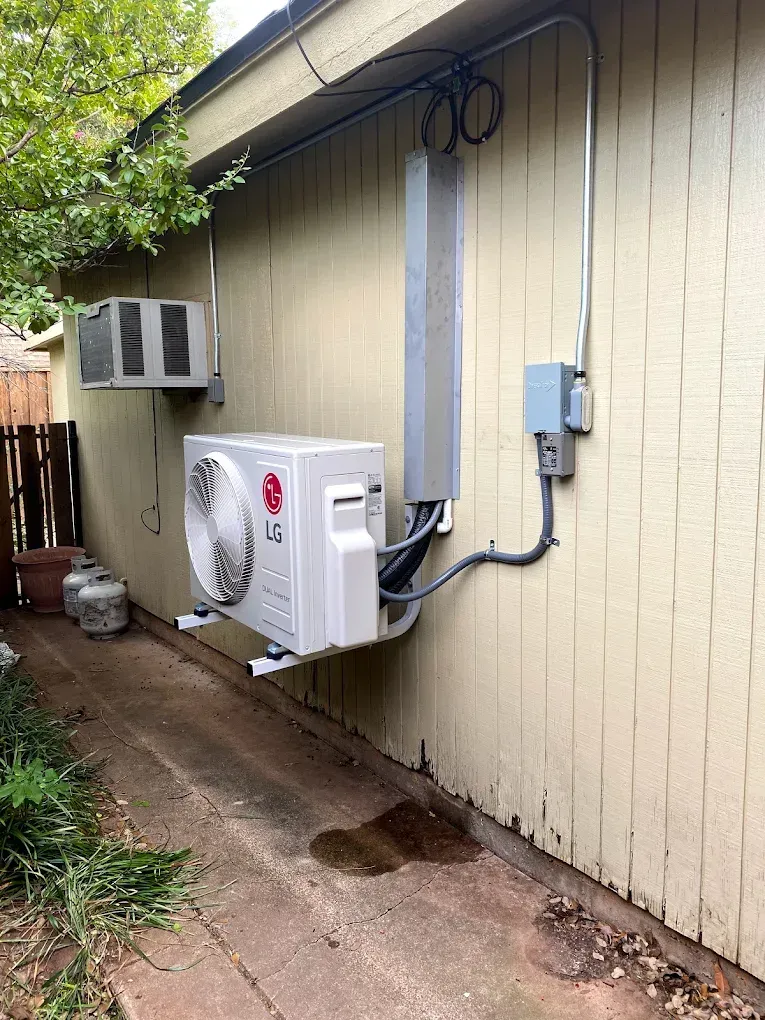 Outdoor AC units mounted on a beige house wall beside a narrow dirt path and electrical conduit.