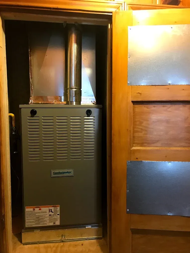 Furnace unit in a narrow wooden utility closet with metal vent pipes and a door partly open.