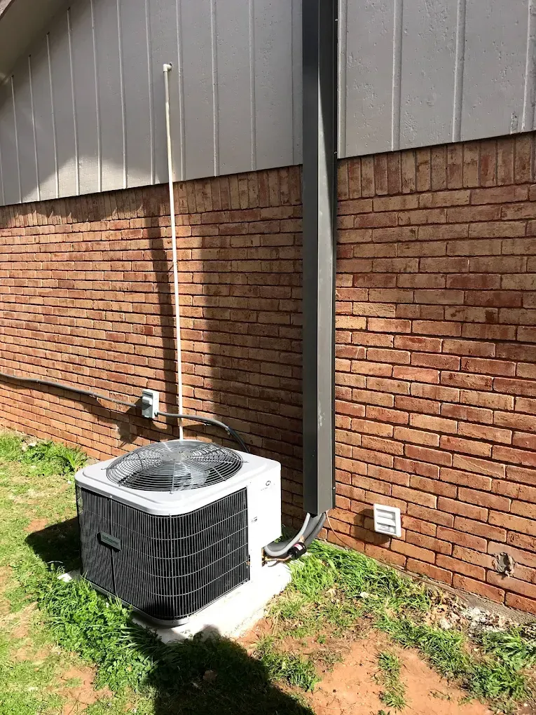 Outdoor air conditioner unit beside a brick wall, with a downspout and house siding above it.