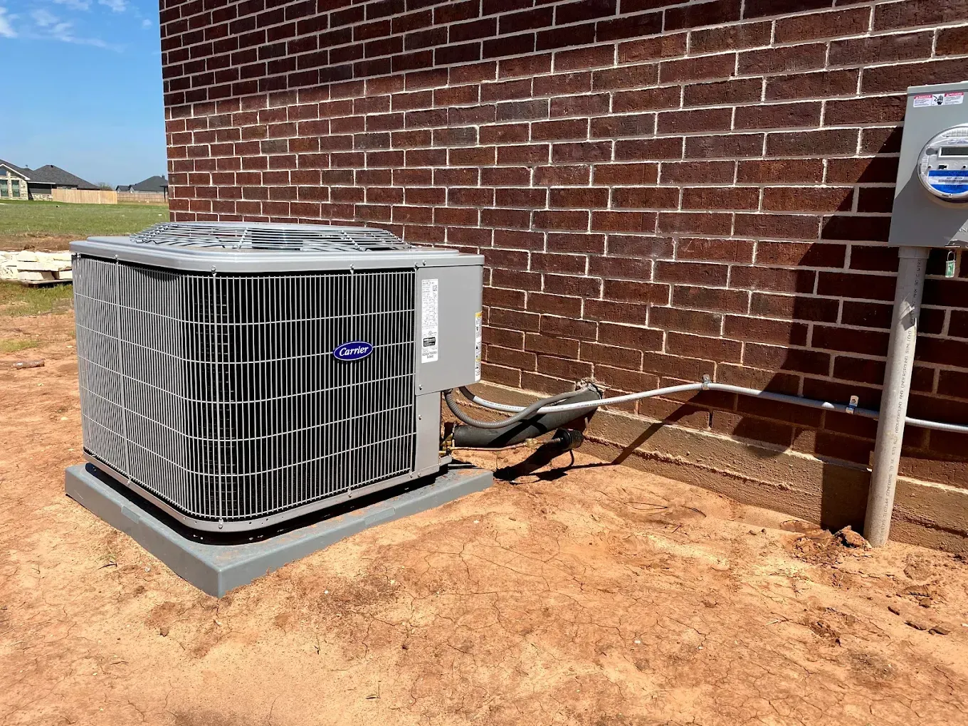 Outdoor HVAC unit beside a brick wall on a dirt lot, with a service line connected to a metal post.