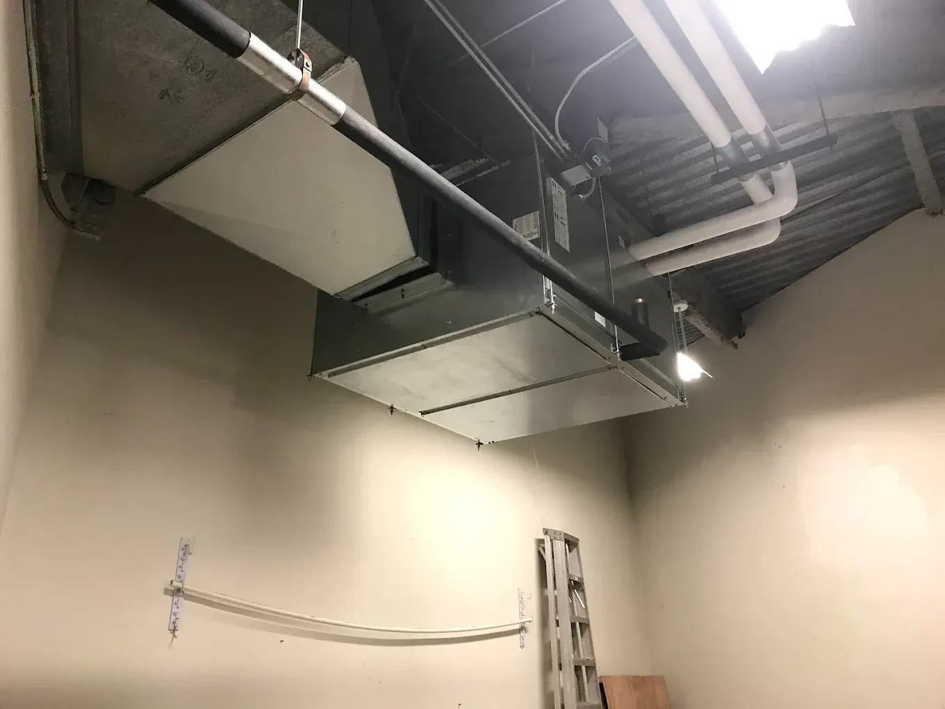 Ceiling-mounted HVAC ducting and pipes in an industrial room with beige walls and fluorescent lighting