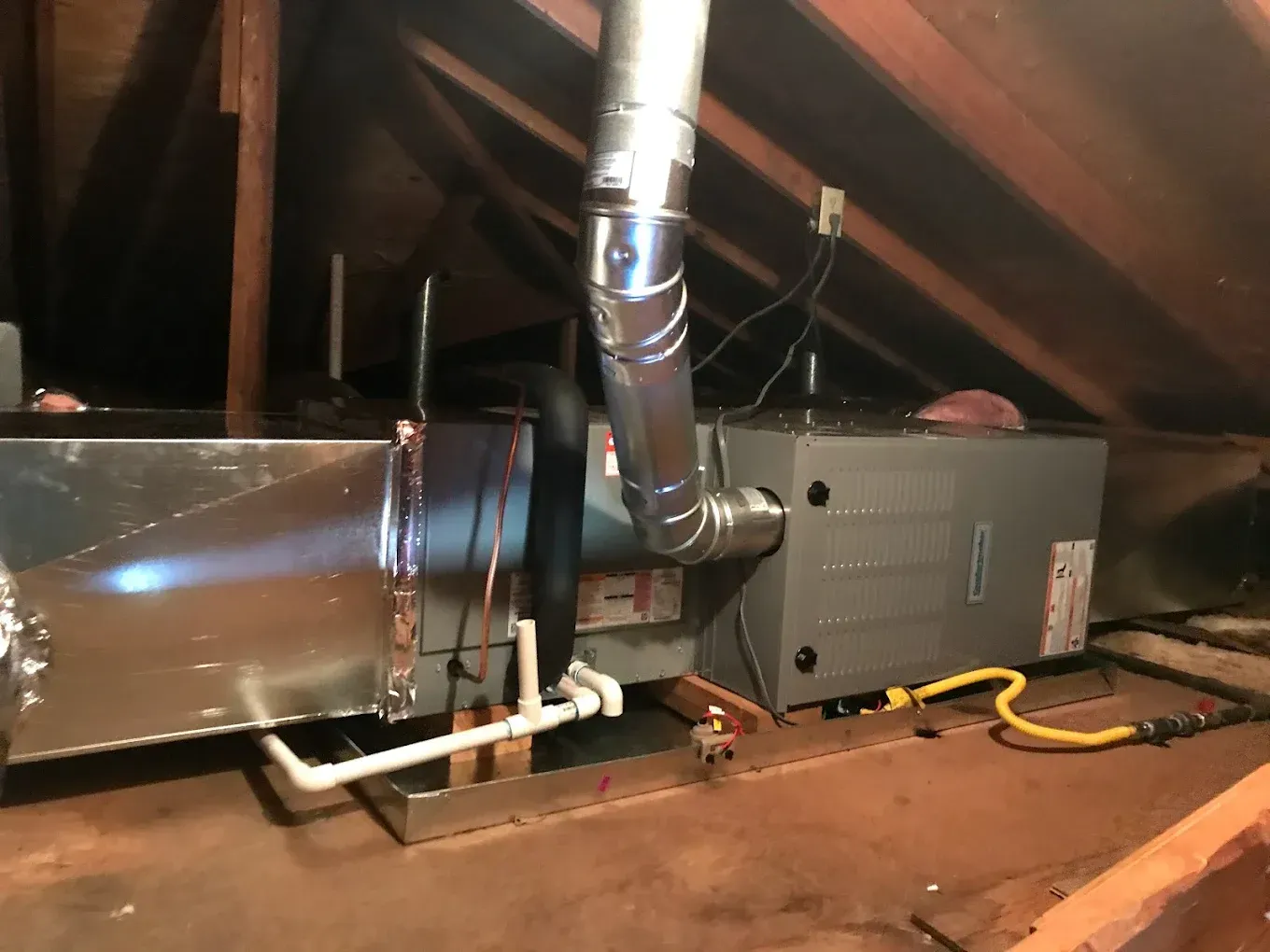 Attic HVAC unit with shiny ductwork and insulated furnace on the floor under exposed rafters