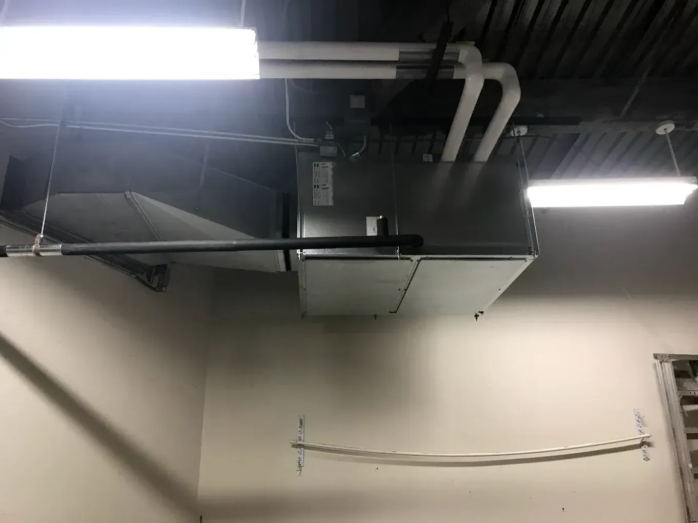 Industrial ceiling with exposed HVAC ducts, bright fluorescent lights, and a wall-mounted cable.
