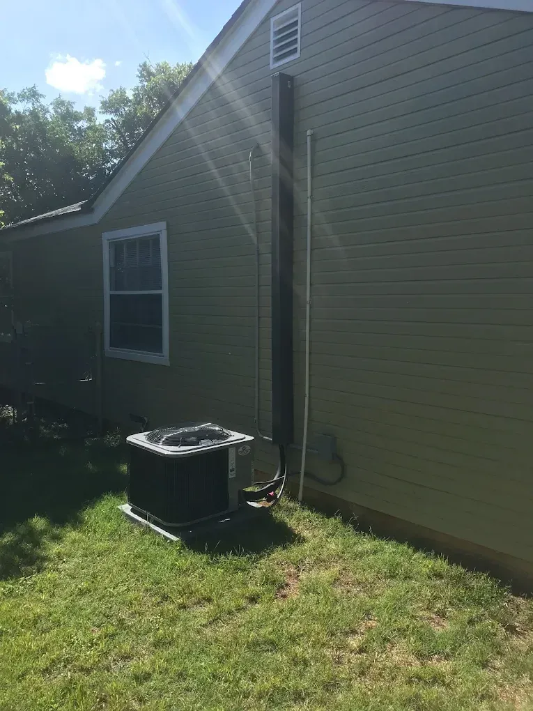 Side of a house with a black outdoor unit, window, and sunlight on the siding and grass.