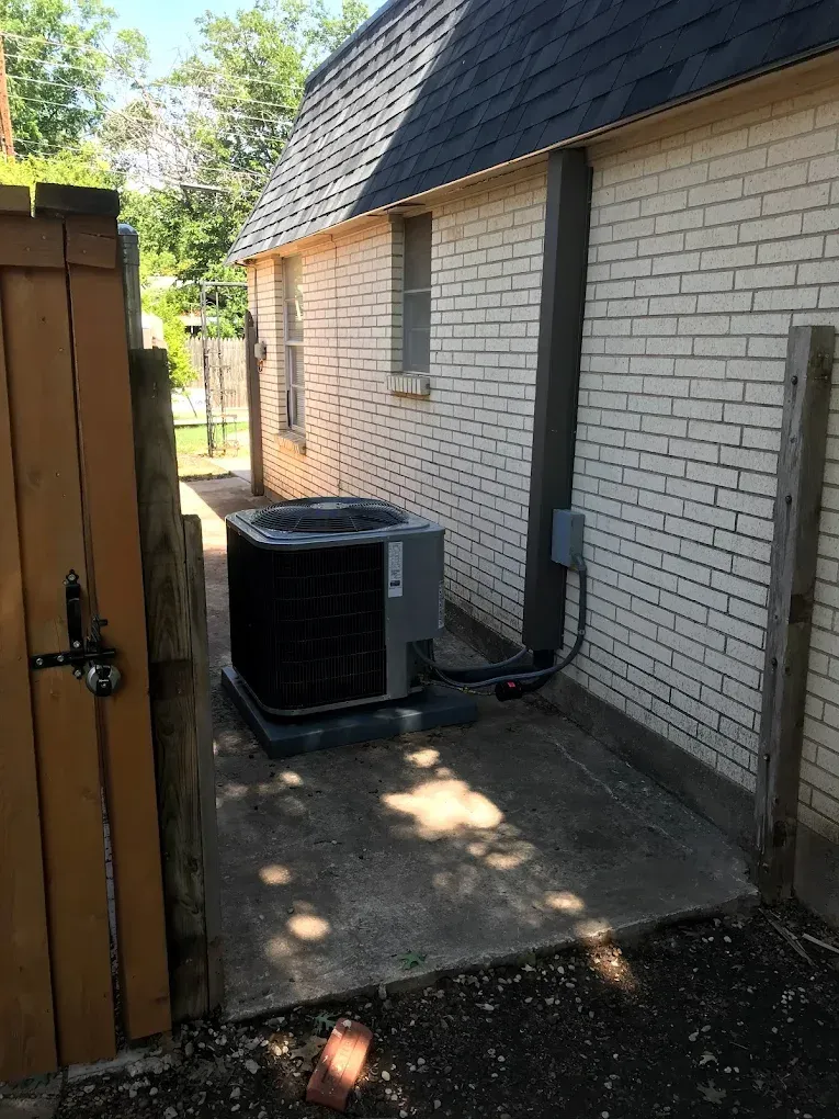 Side yard beside a white brick house with an outdoor AC unit, wooden fence, and paved patio area.