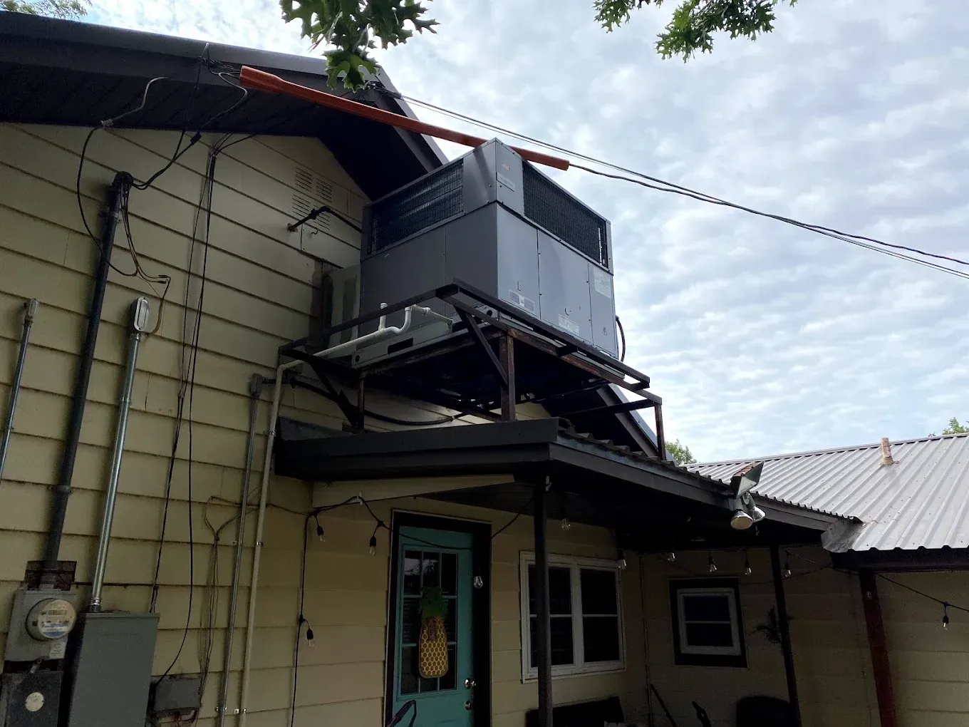 Large HVAC unit mounted above a house entrance with metal supports and wires under a cloudy sky