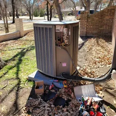 Damaged outdoor air conditioner unit with exposed wiring and debris in a yard ליד brick wall