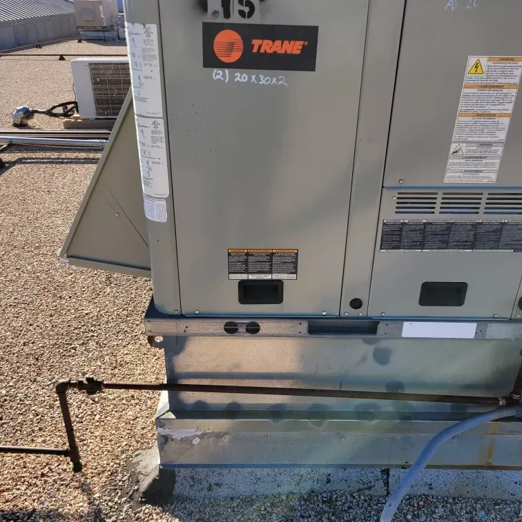 Trane HVAC unit beside a building wall on a gravel rooftop, with pipes and ductwork visible.