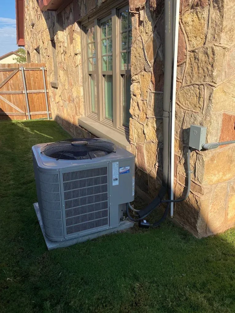 Outdoor air conditioner unit beside a stone house wall and window, on green grass.