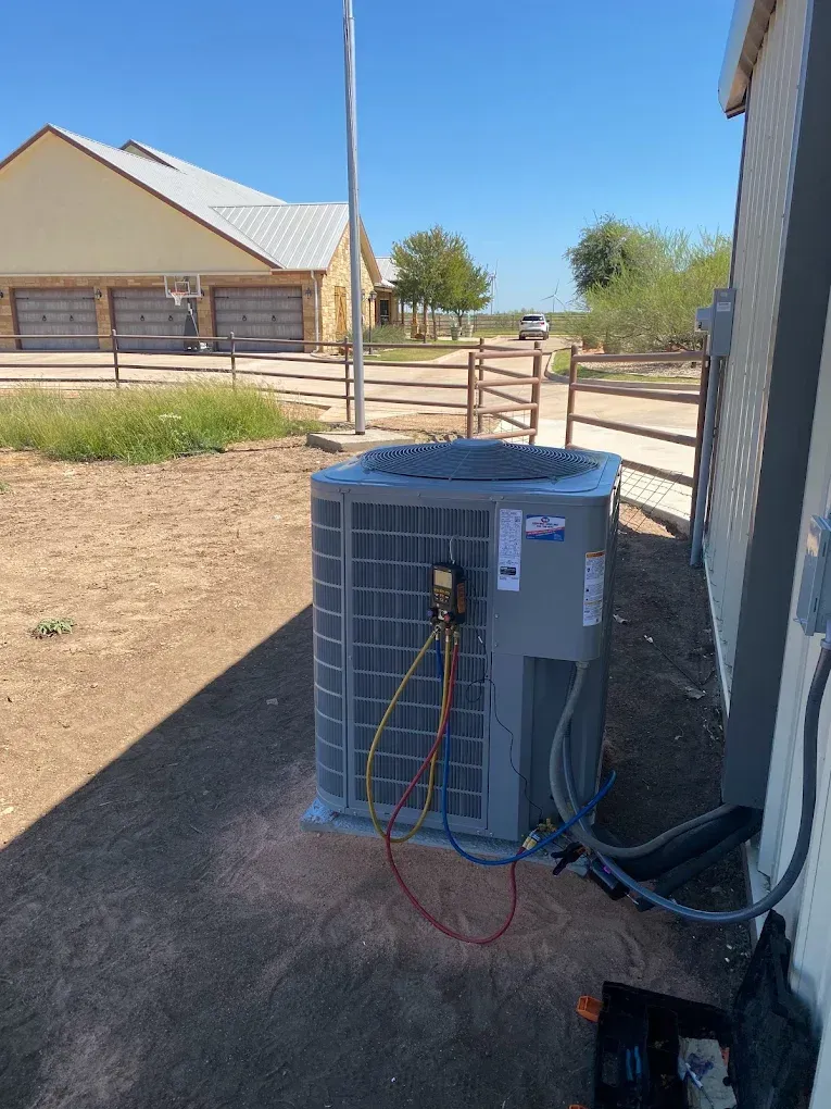 Outdoor HVAC unit beside a building in a dry yard, with exposed wires and a metal fence in the background
