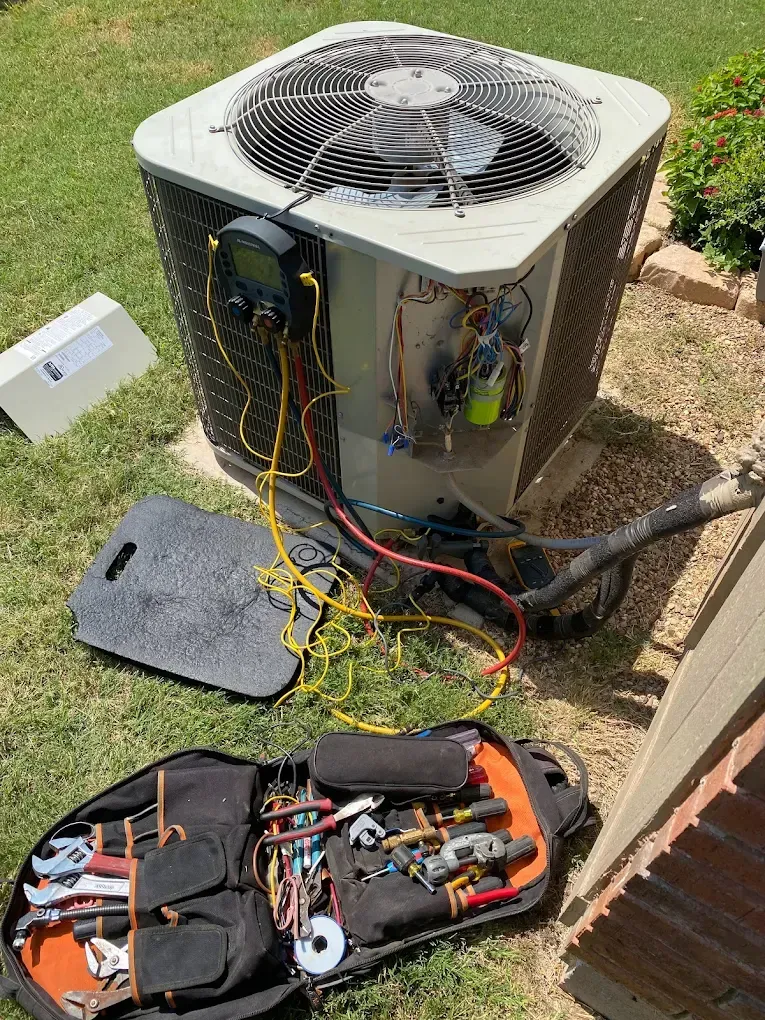 Outdoor air conditioner unit open with tools and wiring spread on the grass nearby