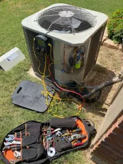 Open HVAC unit with tools and wires on grass beside a brick wall
