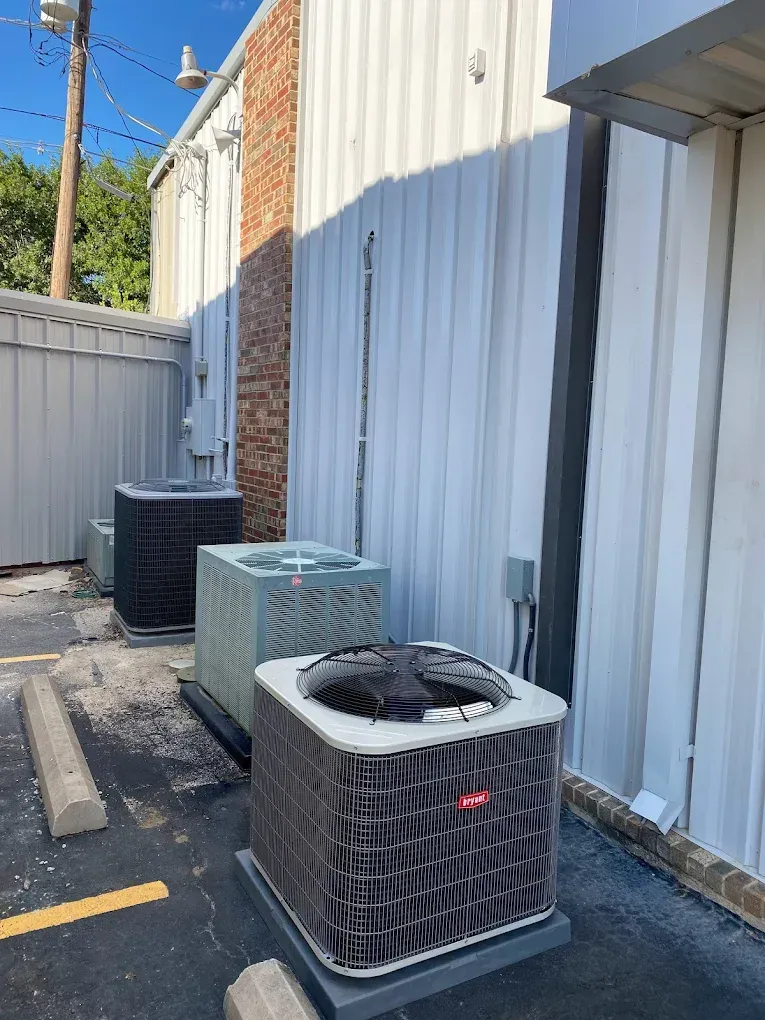 Outdoor HVAC units beside a corrugated metal building, with one unit in the foreground and a brick wall in back.