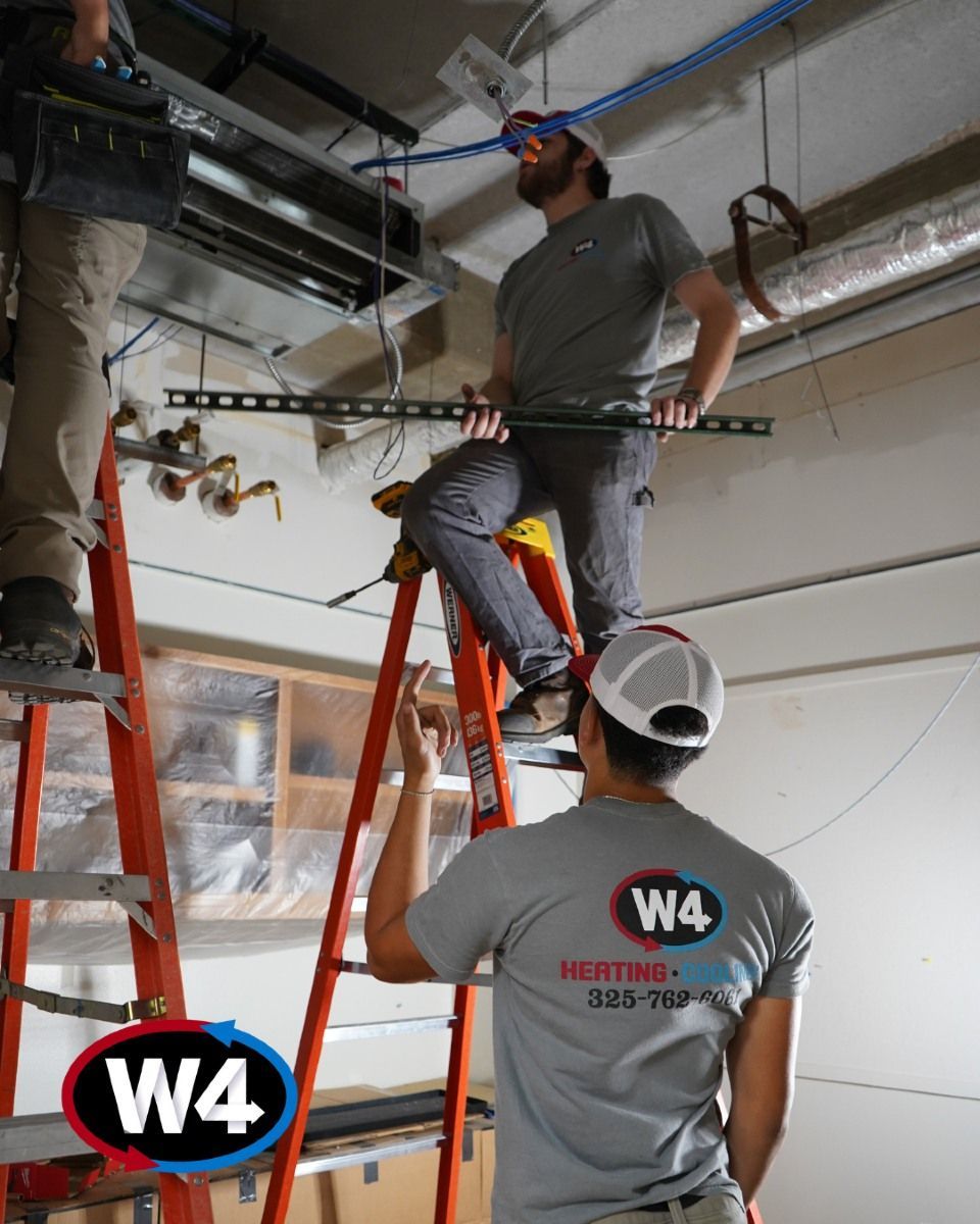Workers on orange ladders inspecting ceiling pipes in an unfinished room with exposed ductwork.