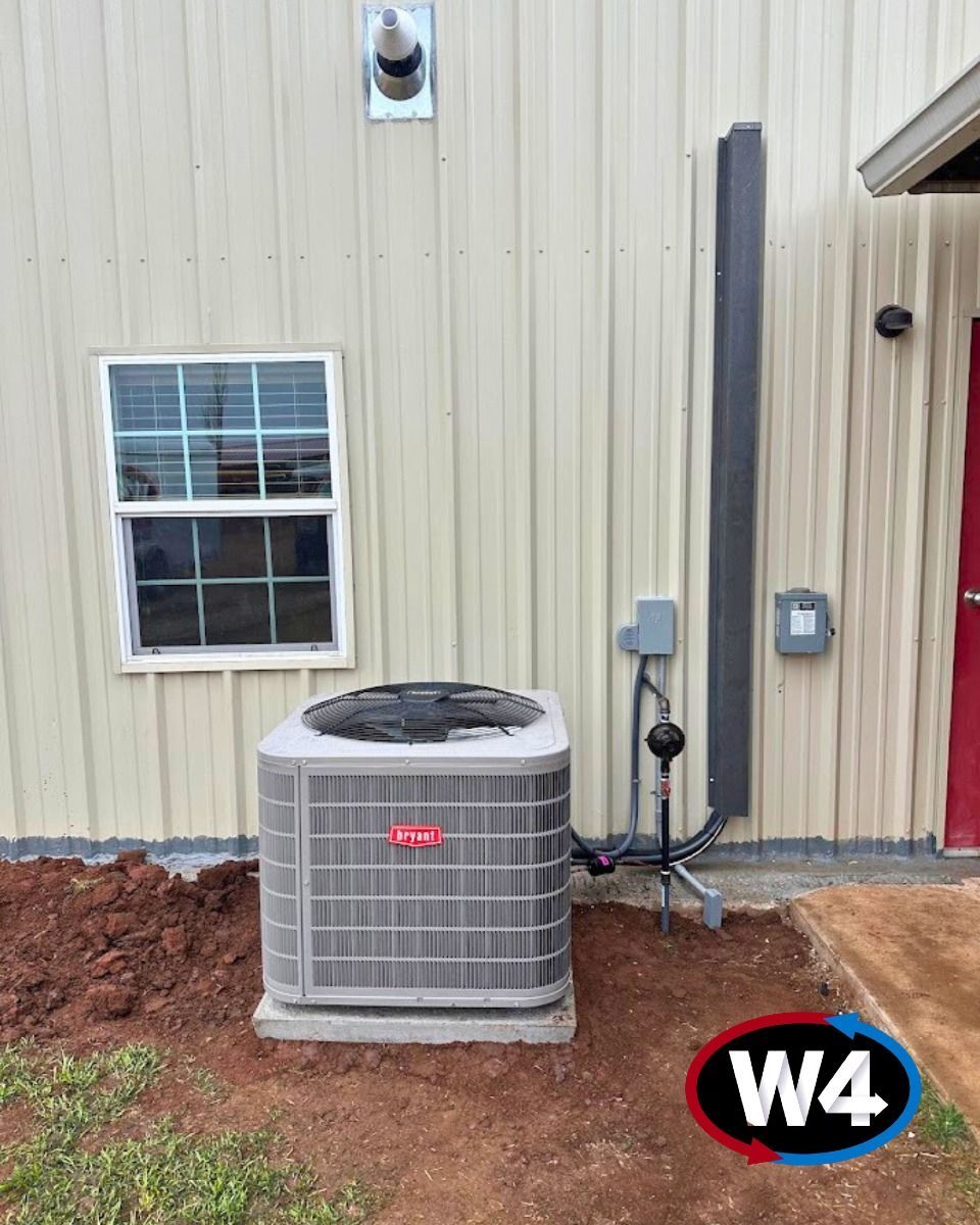 Outdoor HVAC unit beside a tan metal building with a window, utility boxes, and red dirt along the wall.